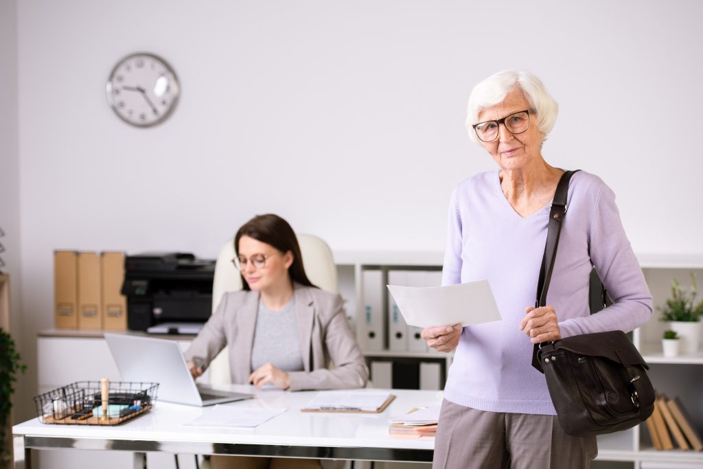 Senior female client of insurance agency with paper and handbag standing in front of camera with agent working on background